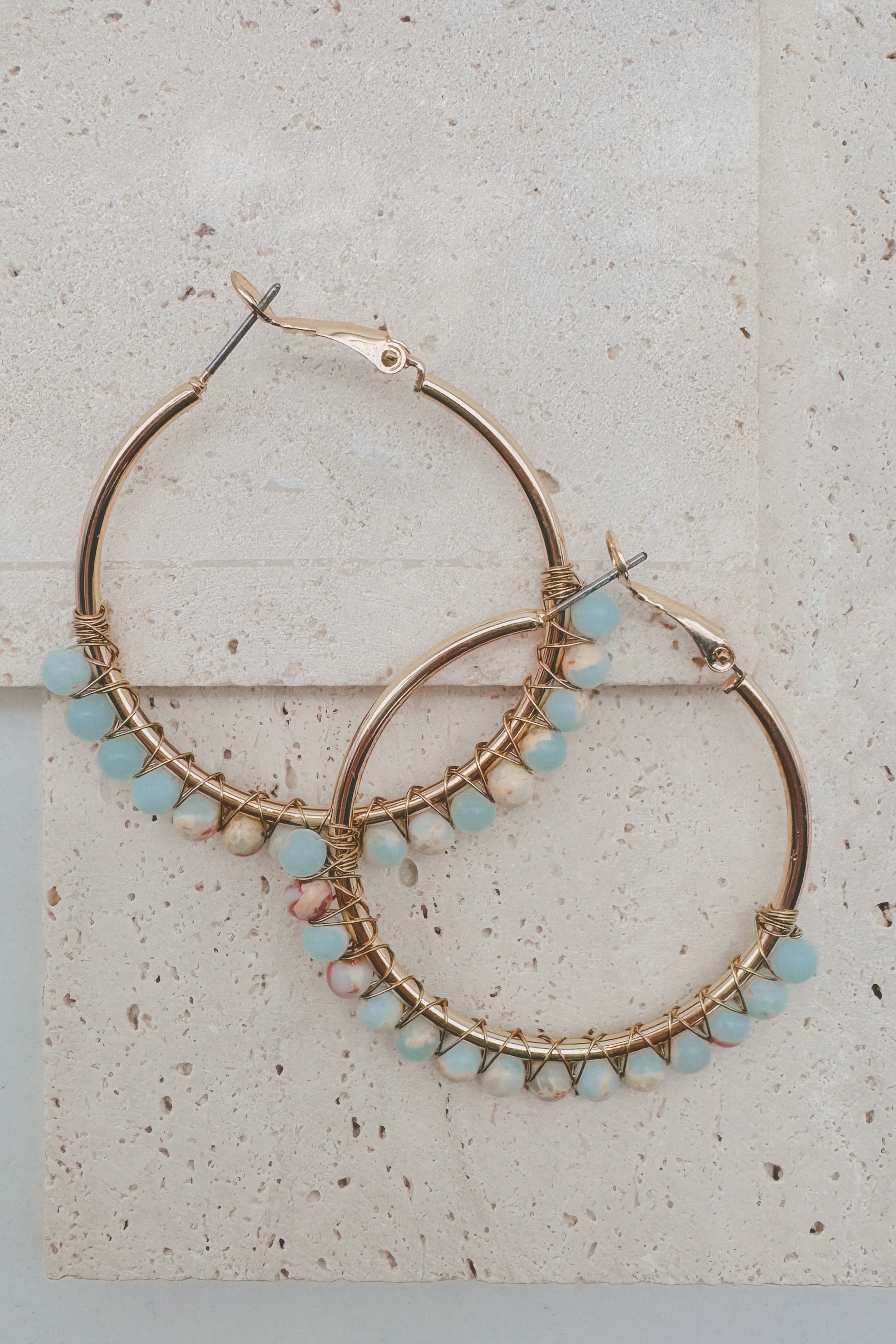 Gold hoop earrings with small light blue, pink, and white beads along the lower edge, displayed on a light beige stone surface.
