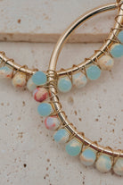 Gold hoop earrings with wrapped light blue and marbled white beads, displayed on a textured beige stone surface.
