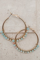 Gold hoop earrings adorned with small light blue and pink beads wrapped in wire, displayed on a light beige stone surface.