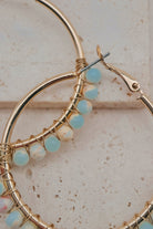 Gold hoop earrings with delicate wire wrapping and small light blue and white beads, shown in a close-up on a textured beige surface.