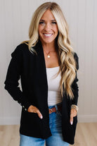 A smiling blonde woman wearing a black open-front blazer over a white tank top and blue jeans, standing against a light paneled wall.
