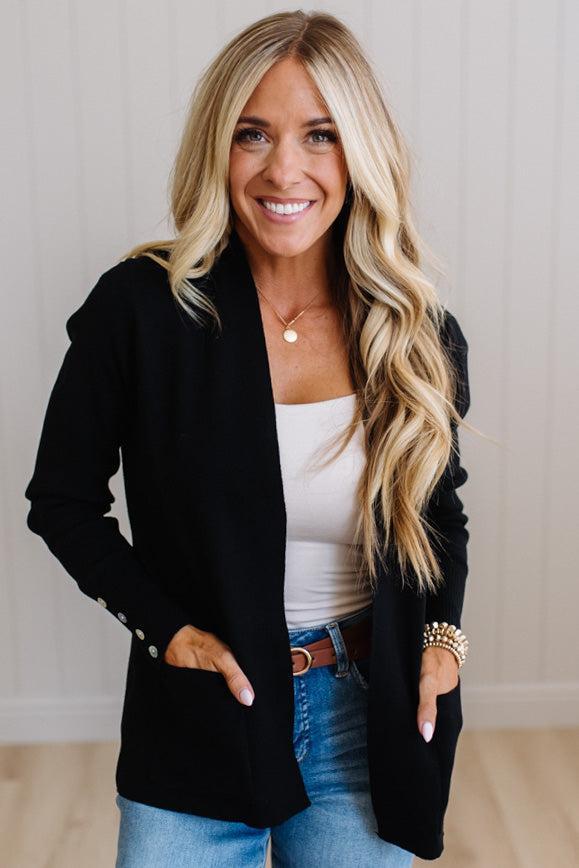 A smiling blonde woman wearing a black open-front blazer over a white tank top and blue jeans, standing against a light paneled wall.