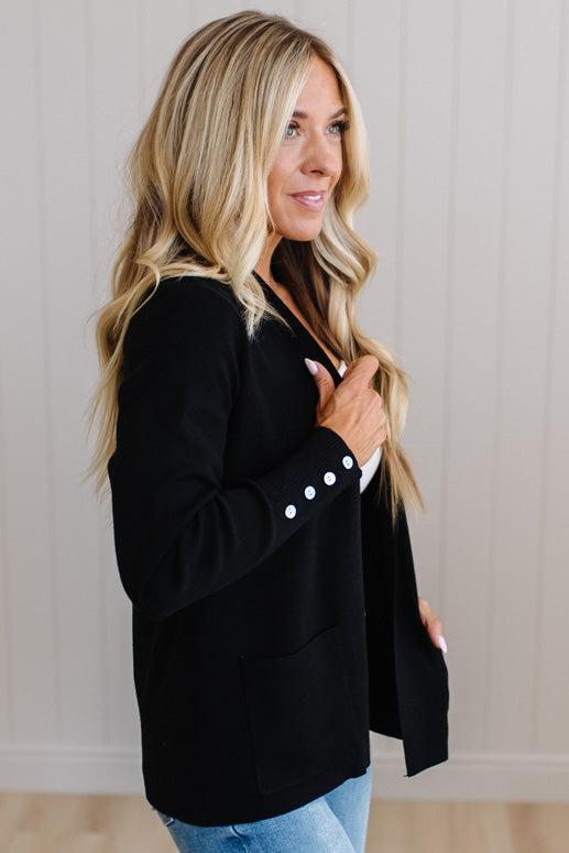 A woman with blonde hair wearing a black long-sleeve blazer with white decorative buttons on the cuffs, paired with light blue jeans, standing against a white paneled wall.