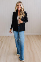 A woman modeling a black longline cardigan with decorative silver buttons on the cuffs, paired with light blue jeans and a beige top, standing in a room with light wood floors and white paneled walls.