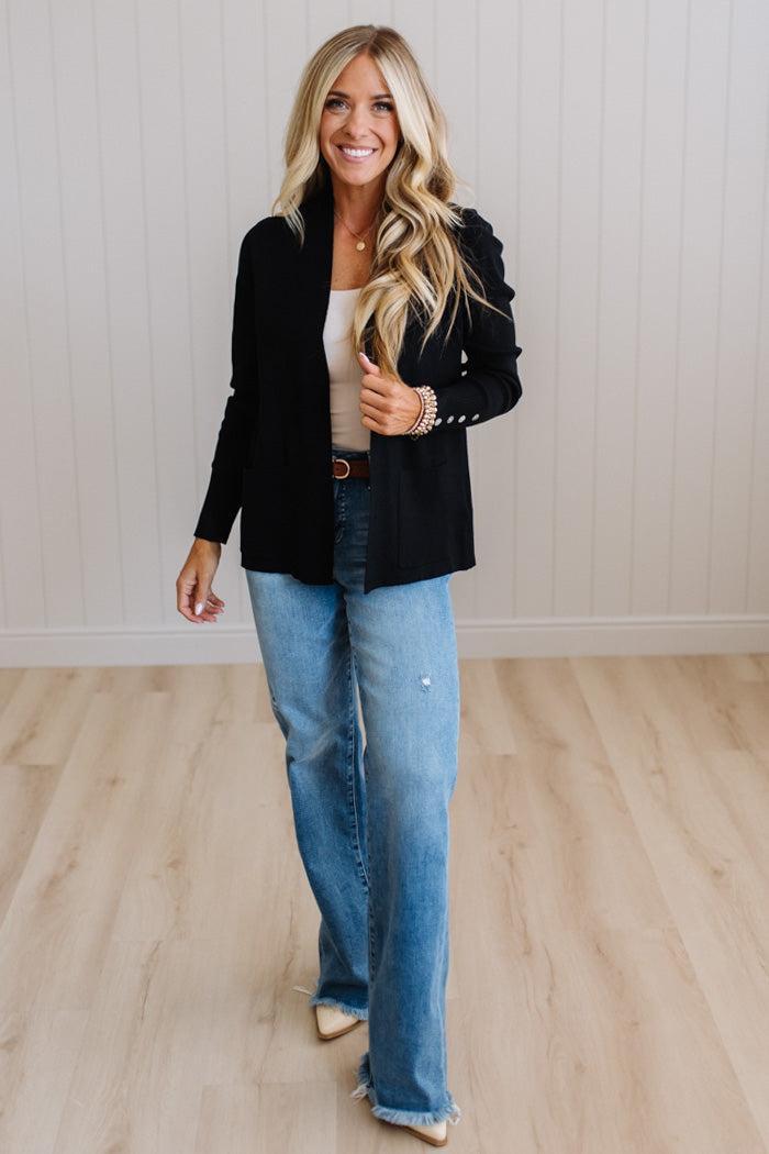 A woman modeling a black longline cardigan with decorative silver buttons on the cuffs, paired with light blue jeans and a beige top, standing in a room with light wood floors and white paneled walls.