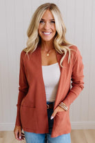 A smiling blonde woman wearing a rust-orange open-front cardigan over a white tank top and blue jeans, standing against a light paneled wall.
