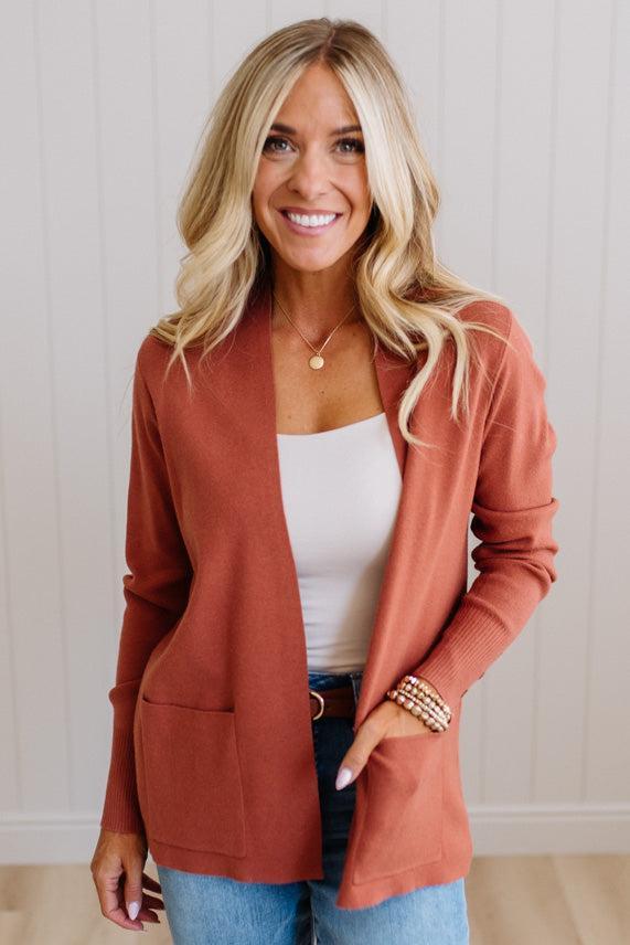 A smiling blonde woman wearing a rust-orange open-front cardigan over a white tank top and blue jeans, standing against a light paneled wall.