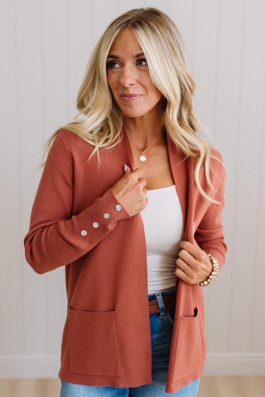 A woman wearing a rust-colored open-front knit cardigan over a white tank top and blue jeans, standing against a light paneled wall.