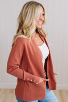 A woman wearing a rust-colored open-front knit cardigan with three decorative buttons on the cuff, styled over a white top and blue jeans, standing against a light wood-paneled wall.