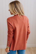 A woman with blonde hair is shown from the back wearing a terracotta-colored knit cardigan with decorative silver buttons on the cuffs, paired with blue jeans, set against a white paneled wall.