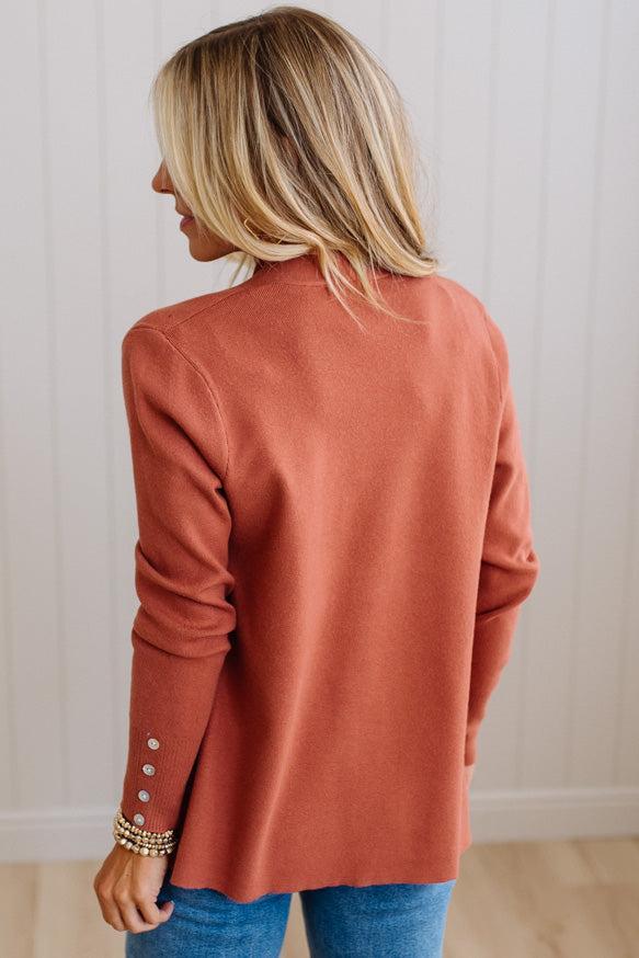 A woman with blonde hair is shown from the back wearing a terracotta-colored knit cardigan with decorative silver buttons on the cuffs, paired with blue jeans, set against a white paneled wall.