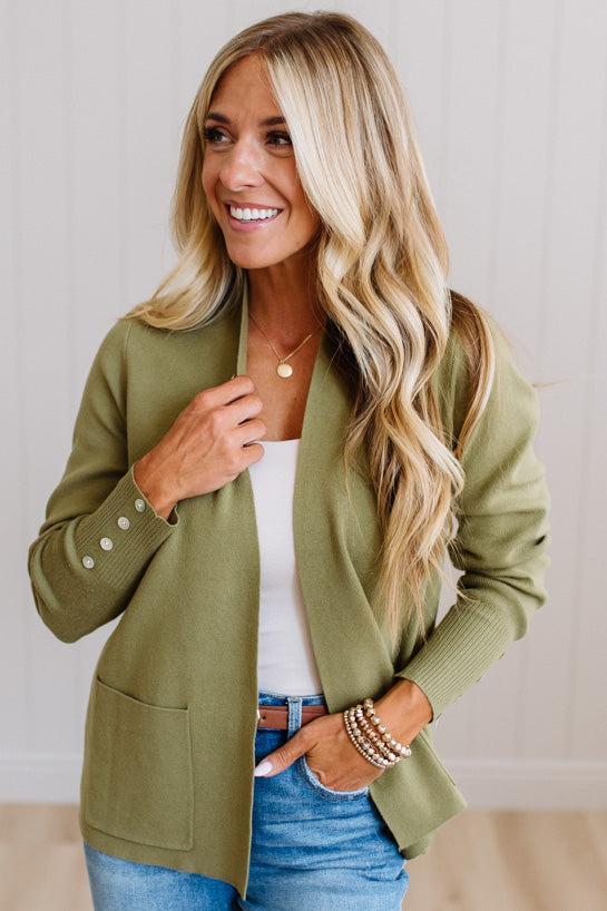 A woman with long blonde hair wearing an olive green knit cardigan over a white top and blue jeans, standing against a light paneled wall.
