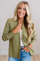 A woman with long blonde hair wearing an olive green knit cardigan over a white top and blue jeans, standing against a light paneled wall.