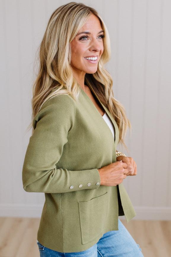 A woman wearing an olive green knit cardigan with button details on the sleeve cuffs, paired with blue jeans, standing against a light wood-paneled wall.