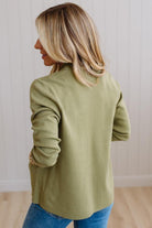 A woman with blonde hair is shown from the back wearing an olive green long-sleeve knit top and blue jeans, standing against a white paneled wall.