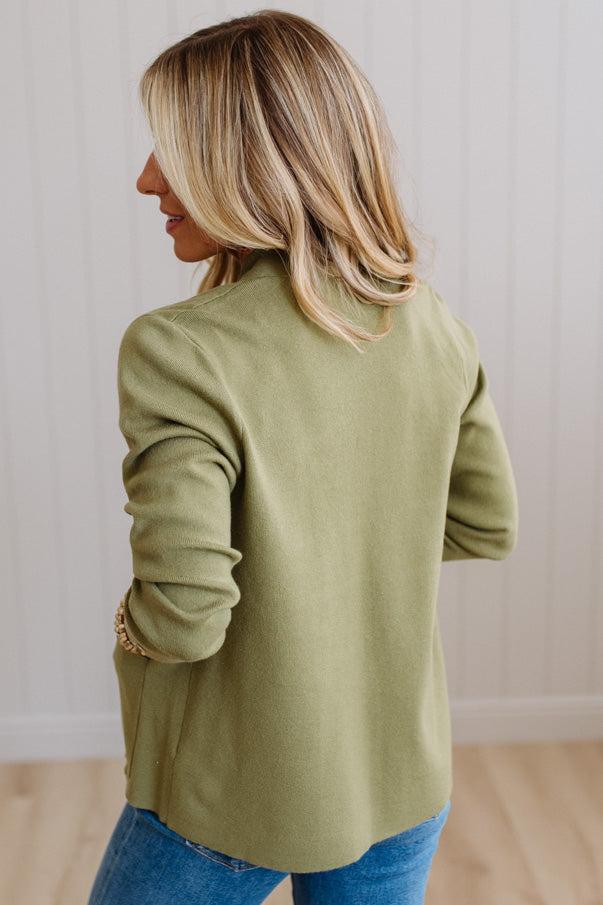 A woman with blonde hair is shown from the back wearing an olive green long-sleeve knit top and blue jeans, standing against a white paneled wall.