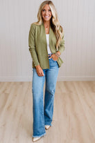 A woman modeling an olive green open-front blazer with decorative buttons, worn over a white top and paired with light-wash blue flare jeans, standing in a room with light wood floors and white paneled walls.