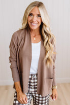 A smiling blonde woman wearing a taupe brown open-front knit cardigan over a white top and brown-and-white gingham pants, standing against a light neutral indoor wall.