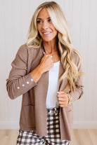 A woman modeling a taupe brown open-front blazer over a white top and brown-and-white plaid pants, standing against a light paneled wall.