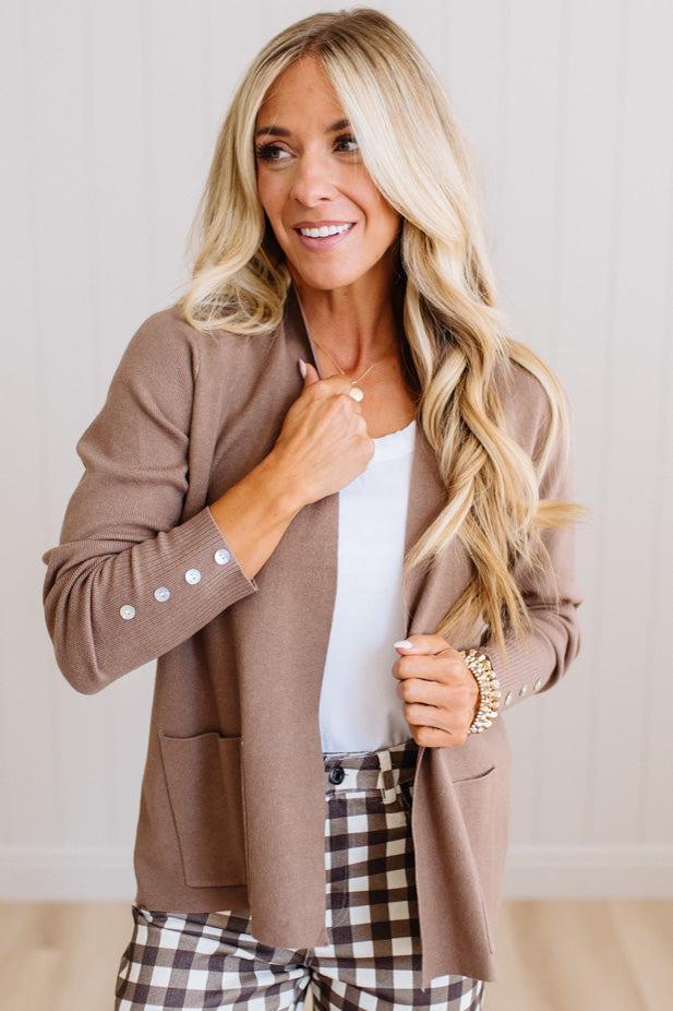 A woman modeling a taupe brown open-front blazer over a white top and brown-and-white plaid pants, standing against a light paneled wall.