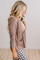A woman wearing a taupe brown knit cardigan with buttoned cuffs and a front pocket, paired with a white top and brown-and-white gingham pants, standing against a white paneled wall.