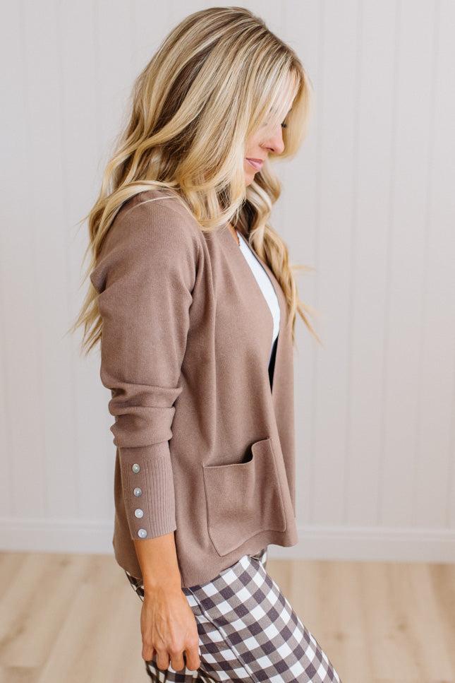 A woman wearing a taupe brown knit cardigan with buttoned cuffs and a front pocket, paired with a white top and brown-and-white gingham pants, standing against a white paneled wall.
