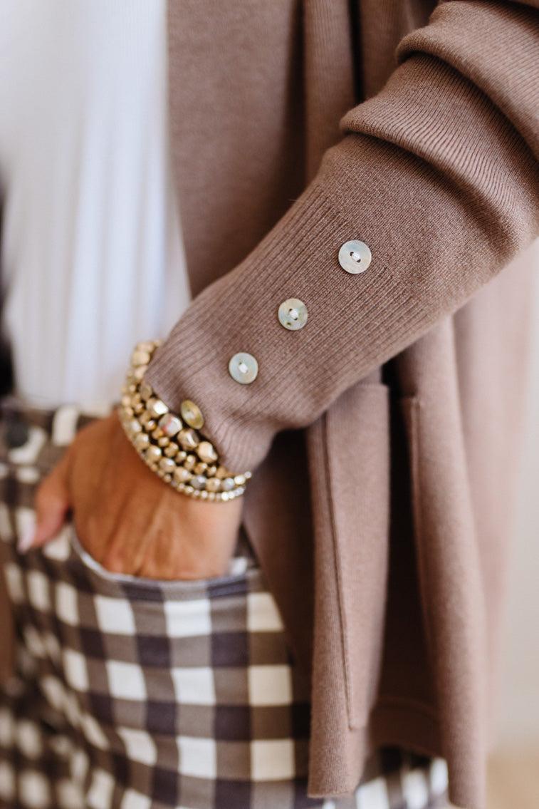 Close-up of a person wearing a taupe knit cardigan with decorative buttons on the sleeve, layered gold beaded bracelets, and a brown-and-white gingham pocket card.