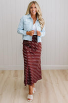 Woman wearing a light blue denim jacket over a white top and burgundy polka dot skirt, standing against a white paneled wall.