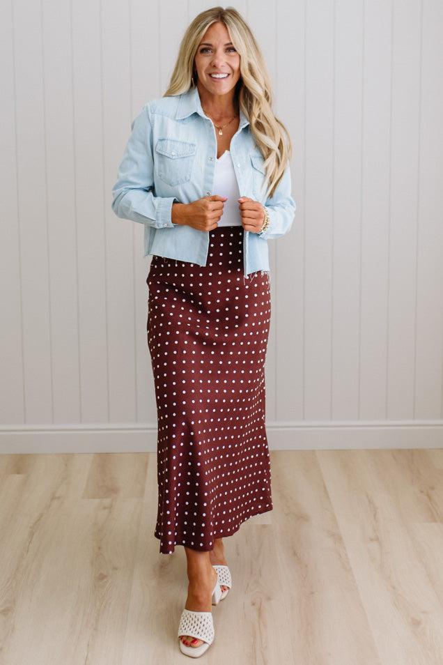 Woman wearing a light blue denim jacket over a white top and burgundy polka dot skirt, standing against a white paneled wall.