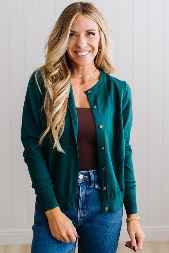 Blonde woman in dark jeans modeling in bright studio wearing a solid green cardigan with brown button front.