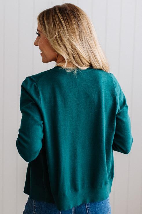 Blonde woman in dark jeans modeling in bright studio wearing a solid green cardigan with brown button front.