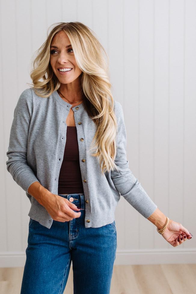 Blonde woman in dark jeans modeling in bright studio wearing a heather grey cardigan with brown button front.
