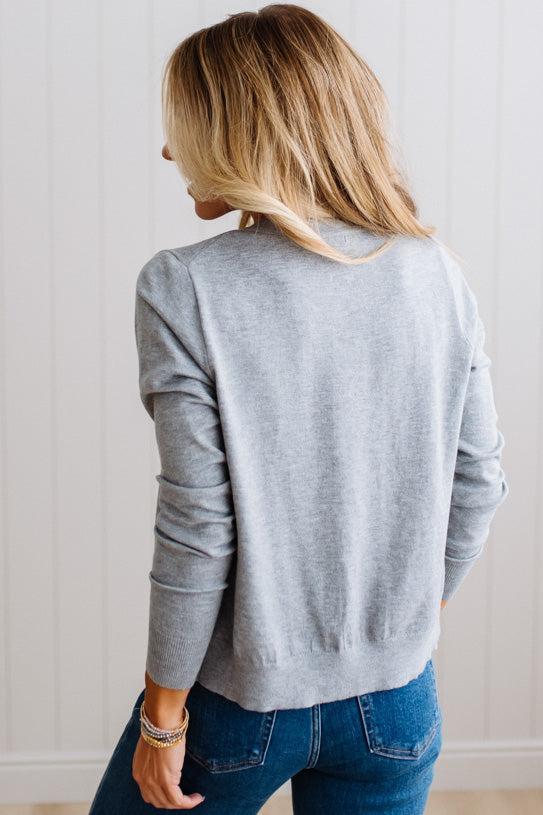 Blonde woman in dark jeans modeling in bright studio wearing a heather grey cardigan with brown button front.