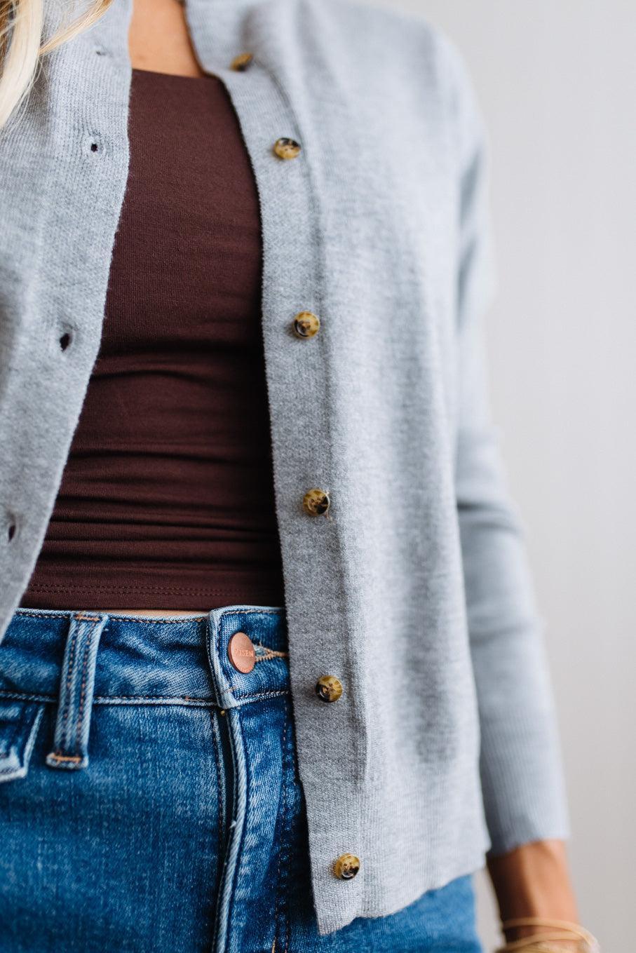 Blonde woman in dark jeans modeling in bright studio wearing a heather grey cardigan with brown button front.