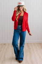 Blonde woman in dark jeans modeling in bright studio wearing a red cardigan with brown button front.