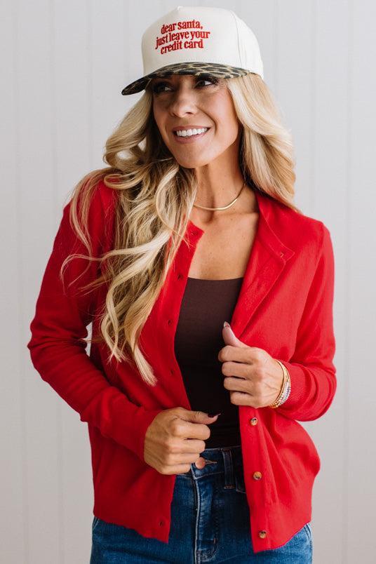 Blonde woman in dark jeans modeling in bright studio wearing a red cardigan with brown button front.