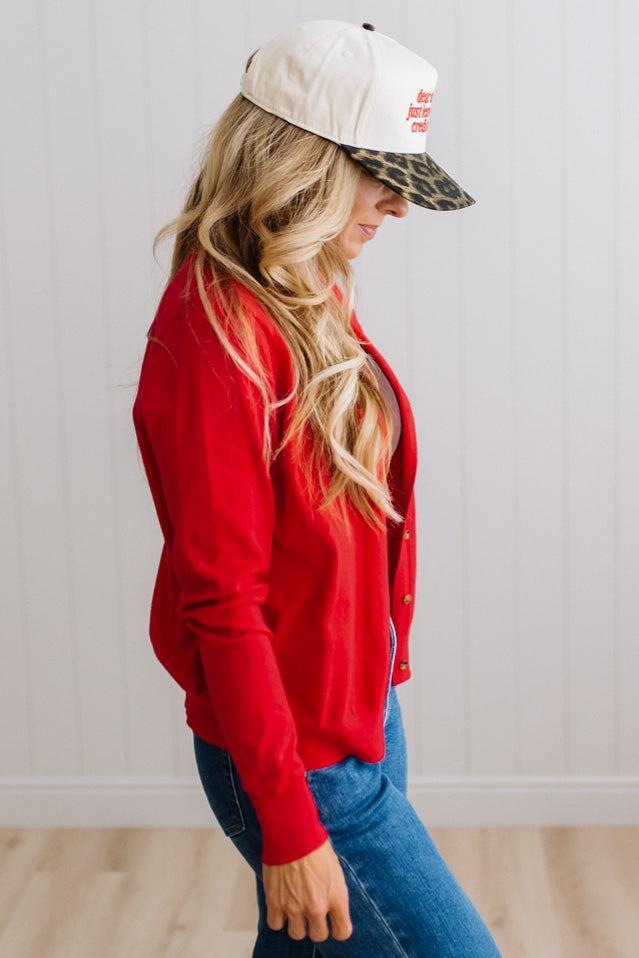 Blonde woman in dark jeans modeling in bright studio wearing a red cardigan with brown button front.
