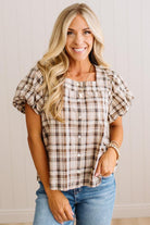 A smiling blonde woman wearing a brown and beige plaid puff-sleeve button-up top with blue jeans, standing against a white paneled wall.