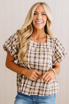 A smiling blonde woman wearing a brown and beige plaid puff-sleeve blouse with blue jeans, standing against a light wood-paneled wall.