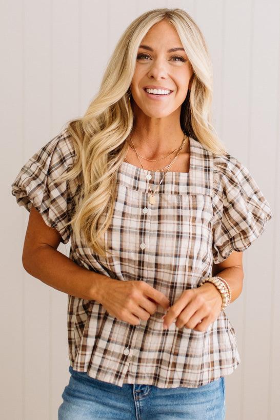 A smiling blonde woman wearing a brown and beige plaid puff-sleeve blouse with blue jeans, standing against a light wood-paneled wall.