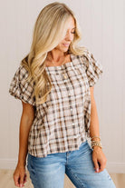 A woman wearing a brown and beige plaid short-sleeve blouse with puff sleeves and a button-down front, paired with blue jeans, standing against a light-colored paneled wall.