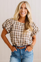 A smiling blonde woman wearing a beige and brown plaid puff-sleeve button-up blouse tucked into blue jeans with a brown belt, standing against a light paneled wall.