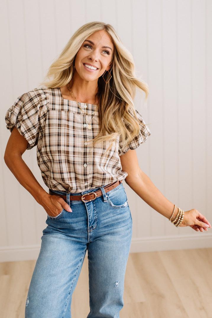 A woman modeling a brown and beige plaid puff-sleeve top with blue jeans and a brown belt, standing in a brightly lit room with white paneled walls and light wood flooring.