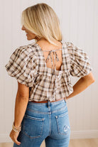 A woman shown from the back wearing a beige and brown plaid puff-sleeve crop top with a tie-back neckline, paired with blue jeans.