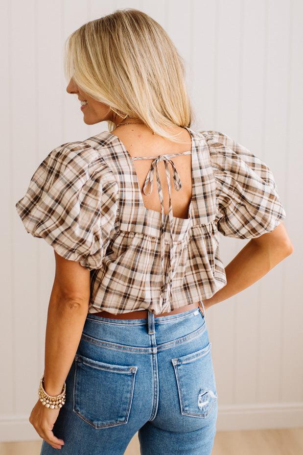 A woman shown from the back wearing a beige and brown plaid puff-sleeve crop top with a tie-back neckline, paired with blue jeans.