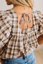 A woman wearing a beige and brown plaid puff-sleeve top with a tie-back neckline, paired with blue jeans.