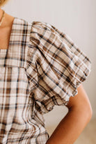 Close-up of a person wearing a beige plaid top with a ruffled puff sleeve and a square neckline.