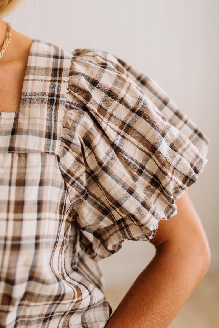 Close-up of a person wearing a beige plaid top with a ruffled puff sleeve and a square neckline.