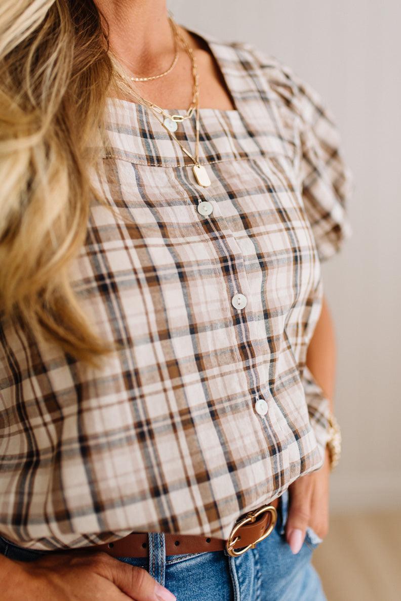 A close-up of a woman wearing a beige and brown plaid puff-sleeve blouse with white buttons, layered gold necklaces, and blue jeans.