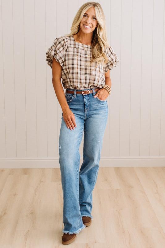 A woman modeling a brown and beige plaid puff-sleeve top with light-wash blue flare jeans, standing in front of a white paneled wall and light wood floor.